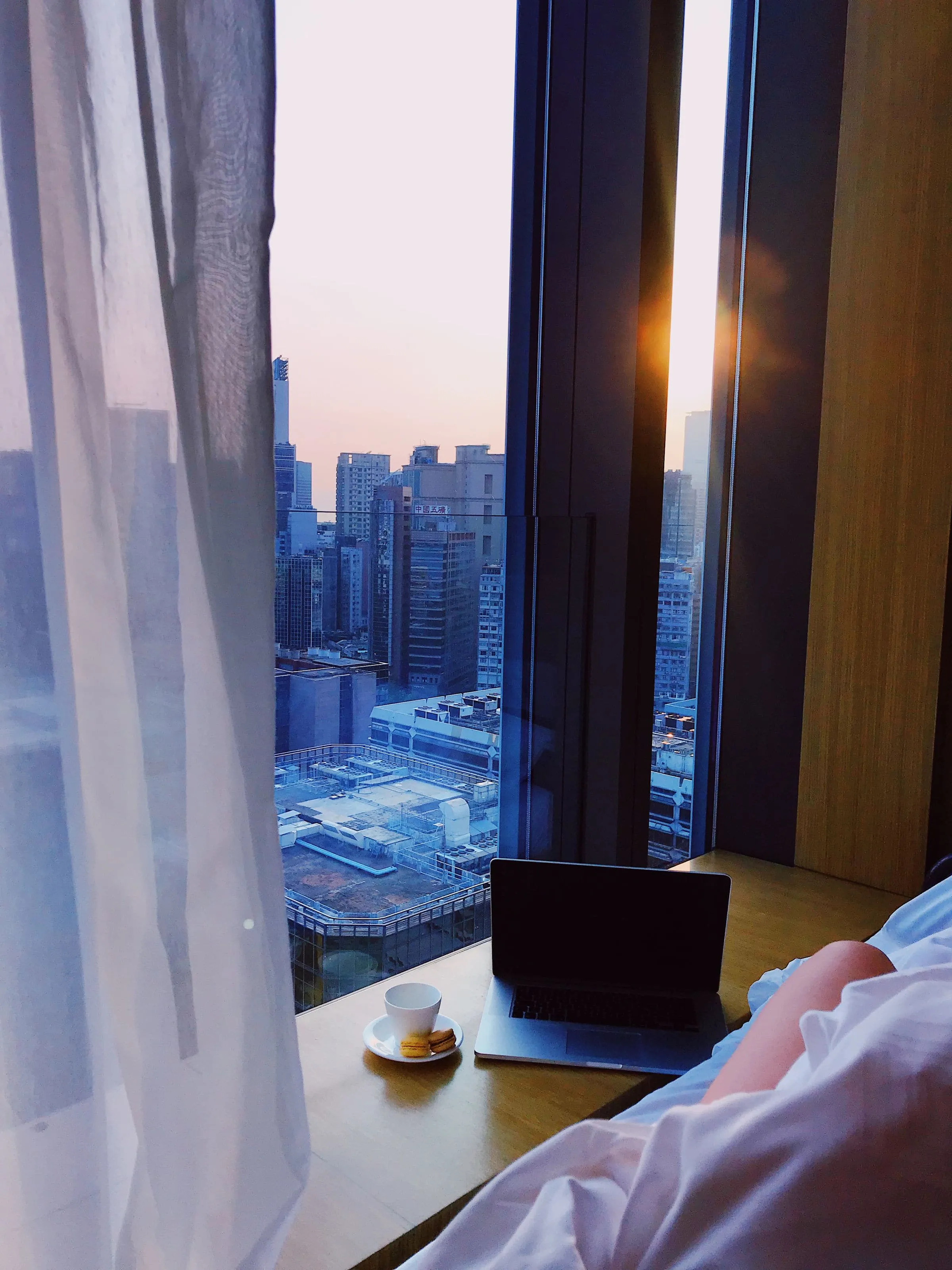 A hotel room window with sheer curtains looking out over a city at dawn