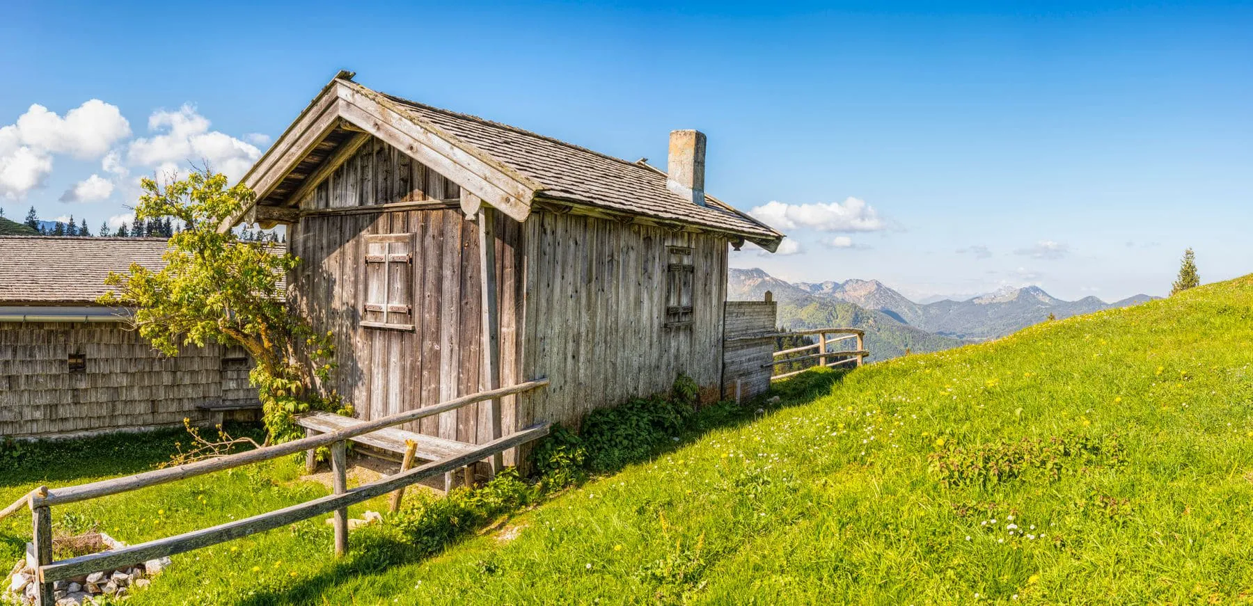 A small alpine hut standing alone on a grassy slope with peaks behind